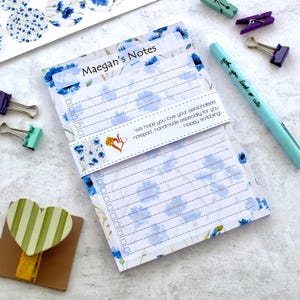 May include: A notepad featuring blue floral designs and the text "Maegan's Notes." The notepad has a checklist and lined pages. A light blue pen with the text "gotcha, you look so cute" is also visible.