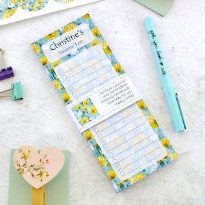 May include: A shopping list notepad with a floral pattern of yellow flowers on a blue backdrop. The notepad is personalized with the name "Christine's SHOPPING LIST". A light blue pen with the text "john, you look so cute" is also visible.