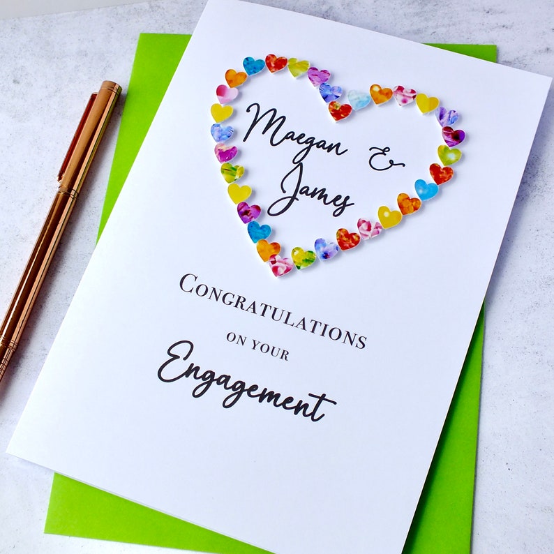May include: A white card with a heart made of colorful paper hearts. The text "Maegan & James" is inside the heart. The text "Congratulations on your Engagement" is below the heart.