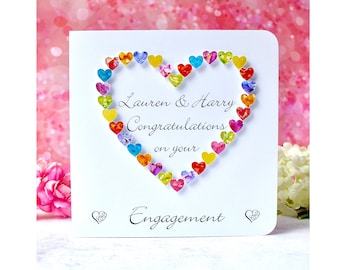 Può includere: Un biglietto bianco con un cuore fatto di cuori di carta colorati. Il biglietto dice "Lauren & Harry Congratulazioni per il vostro fidanzamento".