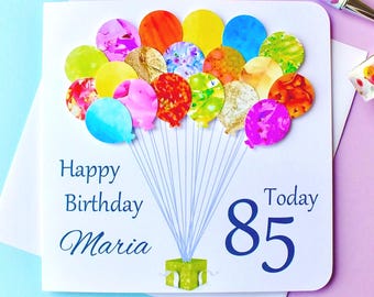 Può includere: Un biglietto bianco con un design di palloncini blu e bianchi. Il biglietto dice "Happy Birthday Maria Today 85".