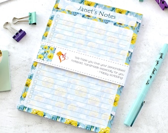 Può includere: Un blocco note con un motivo floreale di fiori gialli su sfondo azzurro. Il blocco note è etichettato "Janet's Notes" e include pagine a righe e un elenco di controllo. È visibile anche una penna azzurra con la scritta "gotke you look so cute".