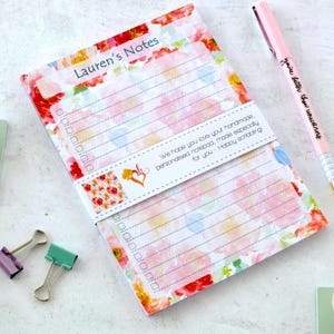 May include: A notepad featuring a pink and white floral design, with the text "Lauren's Notes". A pink pen with the text "You're better than unicorns" is next to the notepad. The notepad is held together with a white band with text.