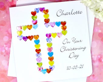 Può includere: Un biglietto bianco con una croce colorata fatta di cuori. Il biglietto dice "Charlotte" e "On Your Christening Day 10-03-21".