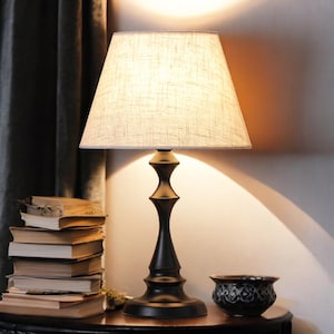 May include: A black table lamp with a beige lampshade, set on a dark wooden table. The lamp has a decorative base and is next to a stack of old books and a small decorative bowl. The lamp is lit, casting a warm glow.