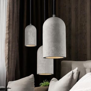 Könnte beinhalten: Drei graue Beton-Pendelleuchten mit zylindrischem Design hängen an schwarzen Kabeln. Die Leuchten strahlen von unten ein warmes Licht aus und schaffen eine moderne und minimalistische Ästhetik. Der Hintergrund zeigt einen dunkelbraunen Vorhang und ein hellgraues Sofa.