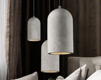 May include: Three gray concrete pendant lights with a cylindrical design hang from black cords. The lights have a warm glow emanating from the bottom, creating a modern and minimalist aesthetic. The background features a dark brown curtain and a light gray sofa.