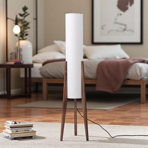May include: A modern floor lamp with a white cylindrical shade and a dark brown wooden tripod base. The lamp is tall, standing on a light-colored rug in a bedroom setting. The lamp's height is approximately 60 inches.