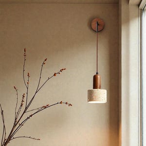 May include: A modern pendant light fixture with a wooden top and a cylindrical stone-like shade. The light hangs from a brown cord attached to a circular wooden mount on the wall. The fixture is illuminated.