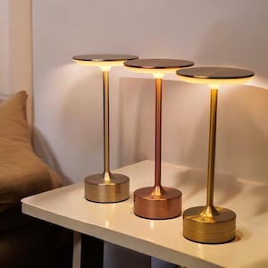 May include: Three modern table lamps with a minimalist design. Each lamp has a circular top and a slender stem, with a round base. The lamps are in gold and copper tones, and are placed on a white surface.