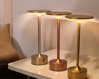 May include: Three modern table lamps with a minimalist design. Each lamp has a circular top and a slender stem, with a round base. The lamps are in gold and copper tones, and are placed on a white surface.