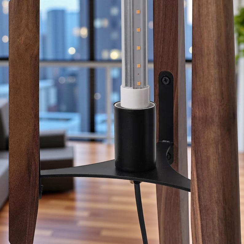 May include: Close-up of a modern floor lamp. The lamp features a black metal base with a triangular support, a black cylindrical lamp holder, and a light-emitting diode (LED) light tube. The lamp has three wooden legs with a dark brown finish.