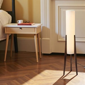 May include: A modern floor lamp with a white cylindrical shade and dark brown wooden legs. A small, light-colored nightstand with a drawer and a book, glasses, and a mug on top sits nearby. The room has a hardwood floor.