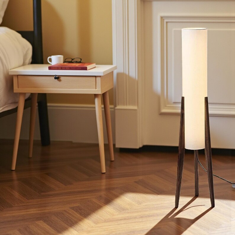 May include: A modern floor lamp with a white cylindrical shade and dark brown wooden legs. A small, light-colored nightstand with a drawer and a book, glasses, and a mug on top sits nearby. The room has a hardwood floor.