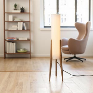 May include: A modern floor lamp with a cylindrical white shade and a wooden tripod base. The lamp is lit, casting a warm glow. The room features a wooden bookshelf, a beige armchair, and a hardwood floor.