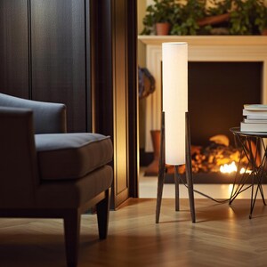 May include: A modern floor lamp with a cylindrical white shade and a dark brown wooden tripod base. The lamp is positioned in a living room setting with a fireplace and armchair in the background. The lamp is approximately 60 inches tall.