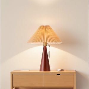May include: A wooden bedside table with a lamp and a mug. The lamp has a pleated shade and a dark red base. A stack of books and a white mug are on the table. The table has two drawers and a shelf.