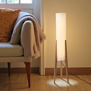 May include: A contemporary floor lamp featuring a rectangular white shade and wooden legs. The lamp is lit, emitting a warm light. The design is minimalist and modern, suitable for living rooms or bedrooms.