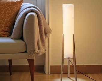 May include: A modern floor lamp with a rectangular white shade and wooden legs. The lamp is illuminated, casting a warm glow. The design is minimalist and contemporary, suitable for living rooms or bedrooms.