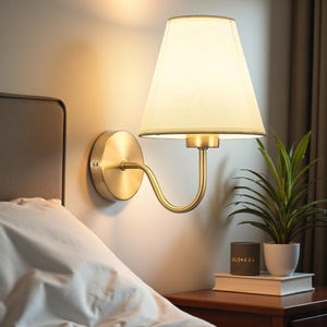 May include: A brass wall sconce with a cream-colored lampshade is mounted on a light-colored wall. The sconce has a curved arm and a circular base. A small potted plant and a candle sit on a wooden nightstand next to a bed.