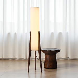 May include: A tall, mid-century modern floor lamp with a cylindrical white shade and dark wood frame. The lamp stands next to a small, round wooden side table. The lamp's height is approximately 60 inches.