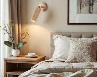 May include: A modern bedroom scene featuring a wooden bedside table with a white vase holding greenery. A beige bed with white pillows and a decorative pillow is visible. A wooden wall-mounted spotlight illuminates the scene.