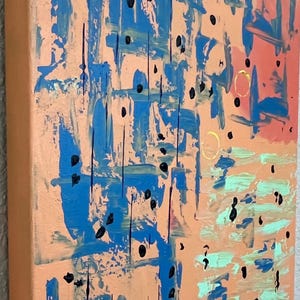 May include: A square canvas painting featuring a peach-coloured background with abstract blue brushstrokes, small black dots, light green horizontal paint marks, and two thin gold circular outlines positioned on the right side of the artwork.