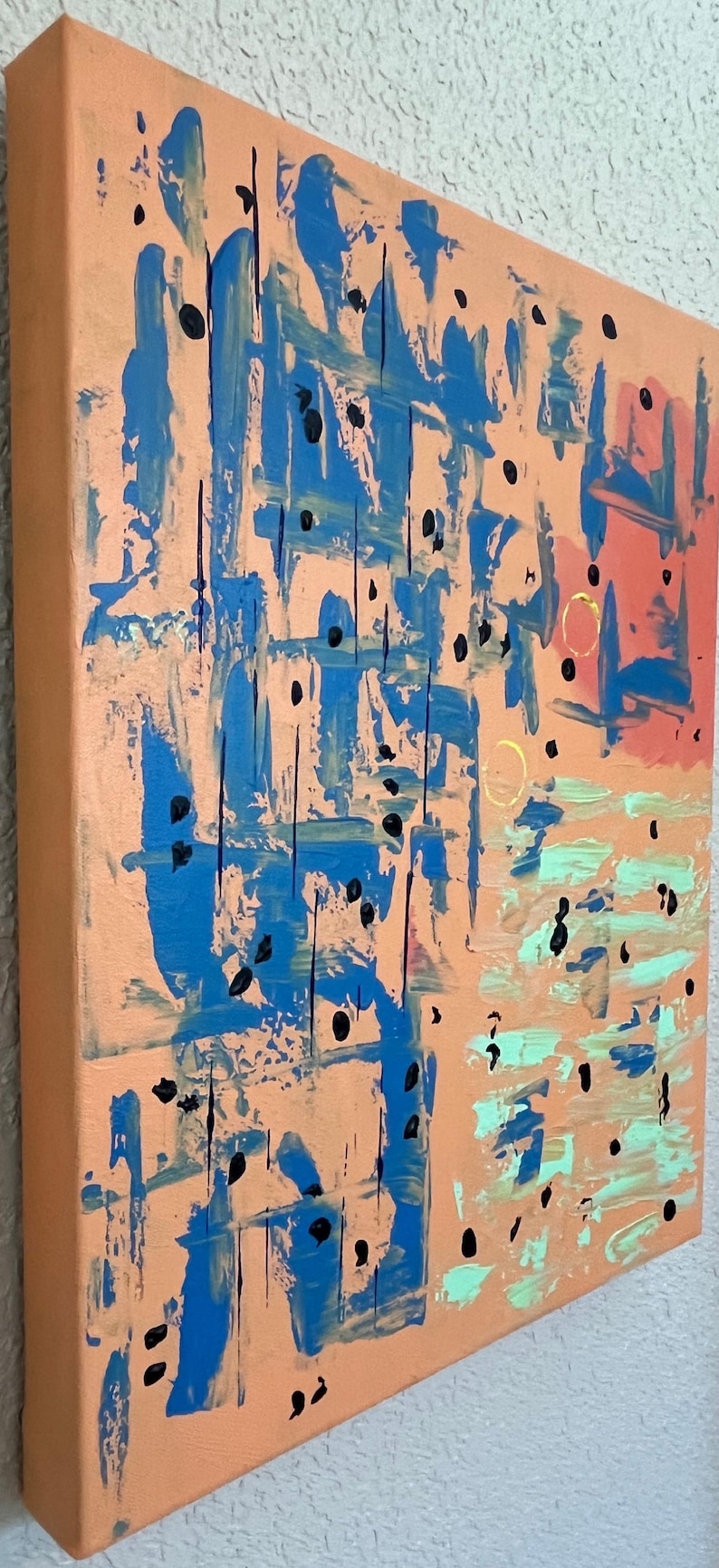 May include: A square canvas painting featuring a peach-coloured background with abstract blue brushstrokes, small black dots, light green horizontal paint marks, and two thin gold circular outlines positioned on the right side of the artwork.
