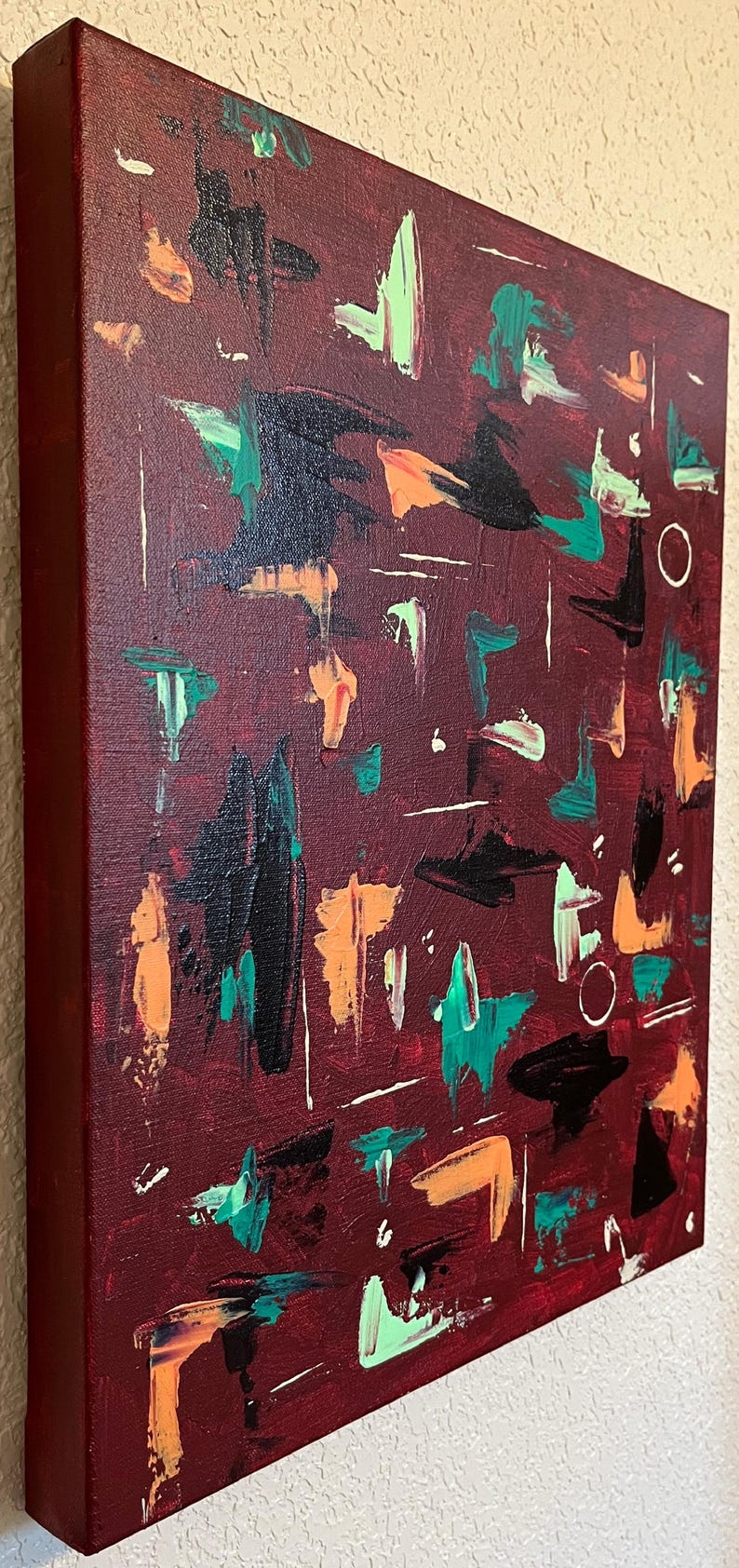 May include: A rectangular canvas painting featuring a deep burgundy background with abstract brushstrokes in black, bright orange, teal, and pale green. The composition includes thin white linear marks and small hand-painted circles scattered across the surface.