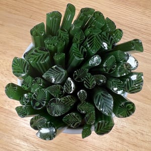 May include: A collection of dark green glass rods, each with a leaf pattern, arranged in a white container. The rods vary in size and shape, creating a textured, organic appearance. The background is a light brown wooden surface.