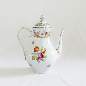 May include: White porcelain coffee pot with a lid, handle, and spout. The pot is decorated with a floral design featuring red, orange, yellow, and purple flowers. Gold trim accents the edges and handle.
