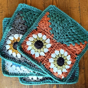 May include: A stack of four handmade crochet squares. Each square features a central flower design with white petals and a dark center, surrounded by orange, teal, and gray yarn. The squares are stacked on top of each other, showcasing the intricate crochet work.