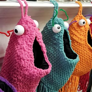 May include: A collection of handmade monster-shaped Christmas stockings in various colours. Each stocking features large googly eyes and pipe cleaner antennae. Colours include purple, pink, teal, orange, red, blue, and lavender.