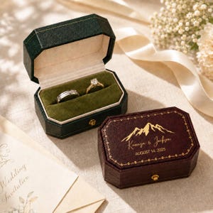 May include: Two octagonal ring boxes, one dark green and open with a silver band and a gold diamond ring inside, and one dark red closed box featuring gold mountain graphics, the text Kamryn & Jackson, and the date AUGUST 14, 2025.