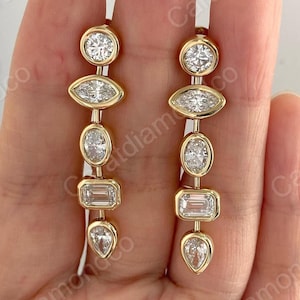 May include: A pair of gold earrings with a series of clear, sparkling gemstones in various shapes. The earrings feature round, marquise, oval, emerald cut, and pear-shaped stones, set in a bezel setting. The earrings are displayed against a neutral background.