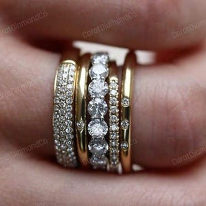 May include: A selection of gold rings set with sparkling diamonds. The rings showcase diverse designs, including bands fully covered in small stones and others with larger, prominent diamonds. The rings are stacked on a finger.