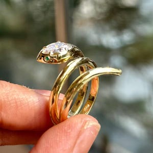 以下が含まれることがあります： 頭に大きな透明な宝石、目に小さな緑色の宝石が付いた、金色のヘビの指輪。 指輪は指に巻き付き、ユニークでエレガントなデザインを作り出しています。 指輪は手で持たれています。