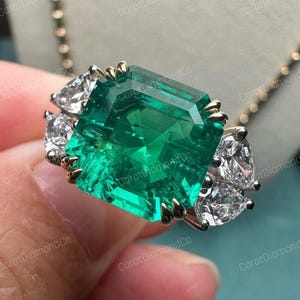 May include: A large emerald-cut green gemstone ring with two pear-shaped white gemstones on each side. The ring has gold and silver accents. The background is a soft blue.
