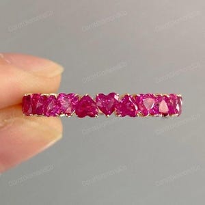 May include: A gold band ring featuring a series of heart-shaped, vibrant pink gemstones. The gemstones are set closely together, creating a continuous row of hearts. The ring is displayed against a soft, neutral background.