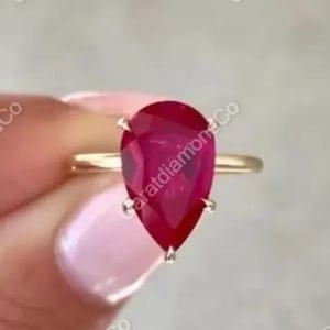May include: A gold ring featuring a large, pear-shaped, deep red gemstone. The ring's band is a thin, polished gold, and the gemstone is held in place by four delicate prongs. The ring is displayed against a soft, neutral background.
