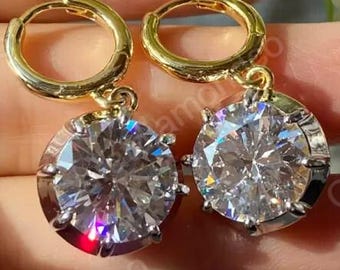 May include: A pair of gold hoop earrings featuring large, round, clear gemstones. The gemstones are faceted, reflecting light and creating a sparkling effect. The earrings are shown against a blurred background.