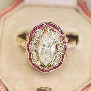 Puede incluir: Un anillo de oro con un diamante central de talla marquesa, rodeado por un halo de pequeños diamantes y piedras preciosas rojas. El anillo, de estilo vintage, se presenta en un joyero rosa.