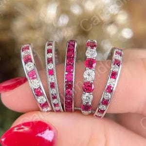 Op de afbeelding: Een collectie van zes zilveren ringen, elk versierd met afwisselende rode edelstenen en heldere, rondgeslepen stenen. De ringen worden op een vinger getoond, waarbij hun ingewikkelde ontwerpen en levendige kleuren worden benadrukt. De edelstenen zijn gezet in een kanaal- of pave-stijl.