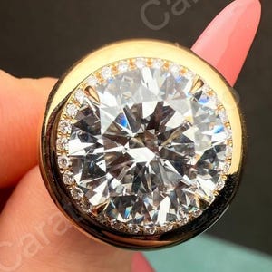 Puede incluir: Un anillo de oro con un gran diamante redondo. El diamante central está rodeado de diamantes más pequeños, engastados en una banda de oro. El diseño del anillo resalta el brillo de los diamantes, creando una pieza lujosa y llamativa.
