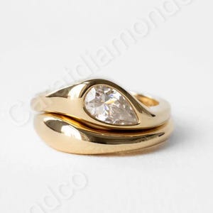 Puede incluir: Dos anillos de oro. El anillo superior presenta un diamante en forma de pera. El anillo inferior es una banda lisa y sencilla. Ambos anillos están sobre un fondo blanco.