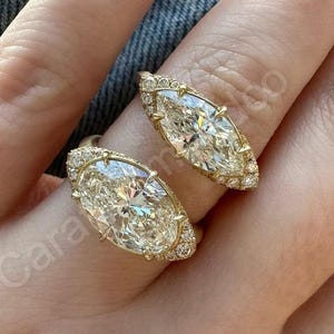 Op de afbeelding: Twee gouden ringen met grote, ovaal geslepen, heldere edelstenen. De ringen zijn gezet met kleinere, heldere stenen rond de hoofdsteen en langs de band. De ringen zitten aan een vinger, met een denim achtergrond.