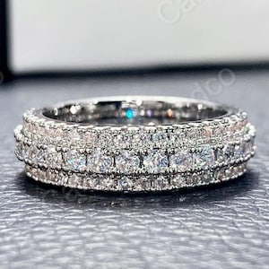 Può includere: Un anello d'argento con più file di piccole pietre preziose trasparenti. L'anello è posto su una superficie scura, con uno sfondo sfocato. Le pietre preziose brillano, riflettendo la luce. L'anello sembra essere uno stile a fascia.
