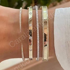 May include: A selection of gold and diamond bracelets. The bracelets include a thin diamond tennis bracelet, a gold bangle with diamonds, and a wide gold bangle. The bracelets are displayed on a wrist.