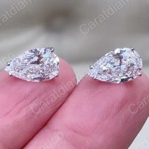 Peut inclure: Deux boucles d'oreilles en diamant en forme de poire. Les diamants sont clairs et étincelants, sertis dans un sertissage en argent ou en or blanc. Les boucles d'oreilles sont présentées sur une surface rose. L'image contient le texte "Caratdiamondco".