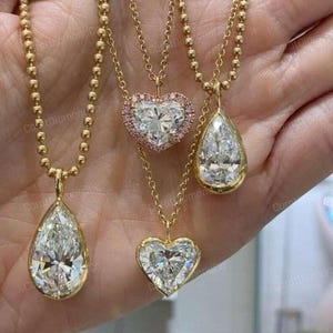 May include: Four gold necklaces with diamond pendants. Two pendants are teardrop-shaped, one is a heart shape, and another is a heart shape surrounded by pink stones. The necklaces are on a gold beaded chain.